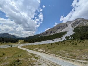 PXL_20260729_121952438 - Wasser & Morphologie Engineering Winding dirt path through a grassy alpine meadow toward a rocky mountain ridge under a blue, partly cloudy sky.