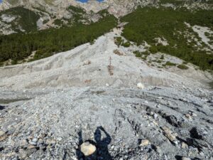 PXL_20260730_080633459 - Wasser & Morphologie Engineering Steep mountain slope covered in loose rocks and scree with evergreen trees on both sides under a clear blue sky.